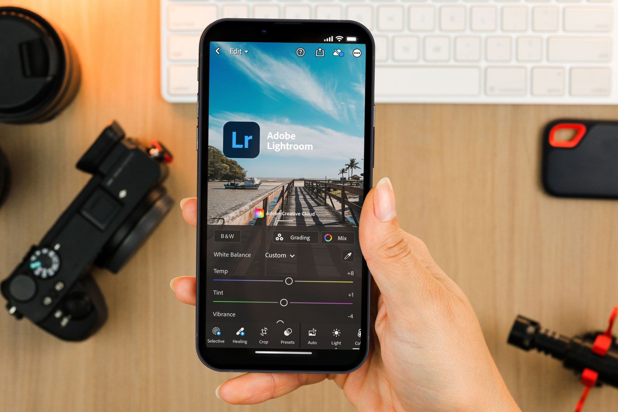 Bàn tay cầm điện thoại thông minh, hiển thị ứng dụng Lightroom Mobile, với thiết bị máy ảnh ở phía sau