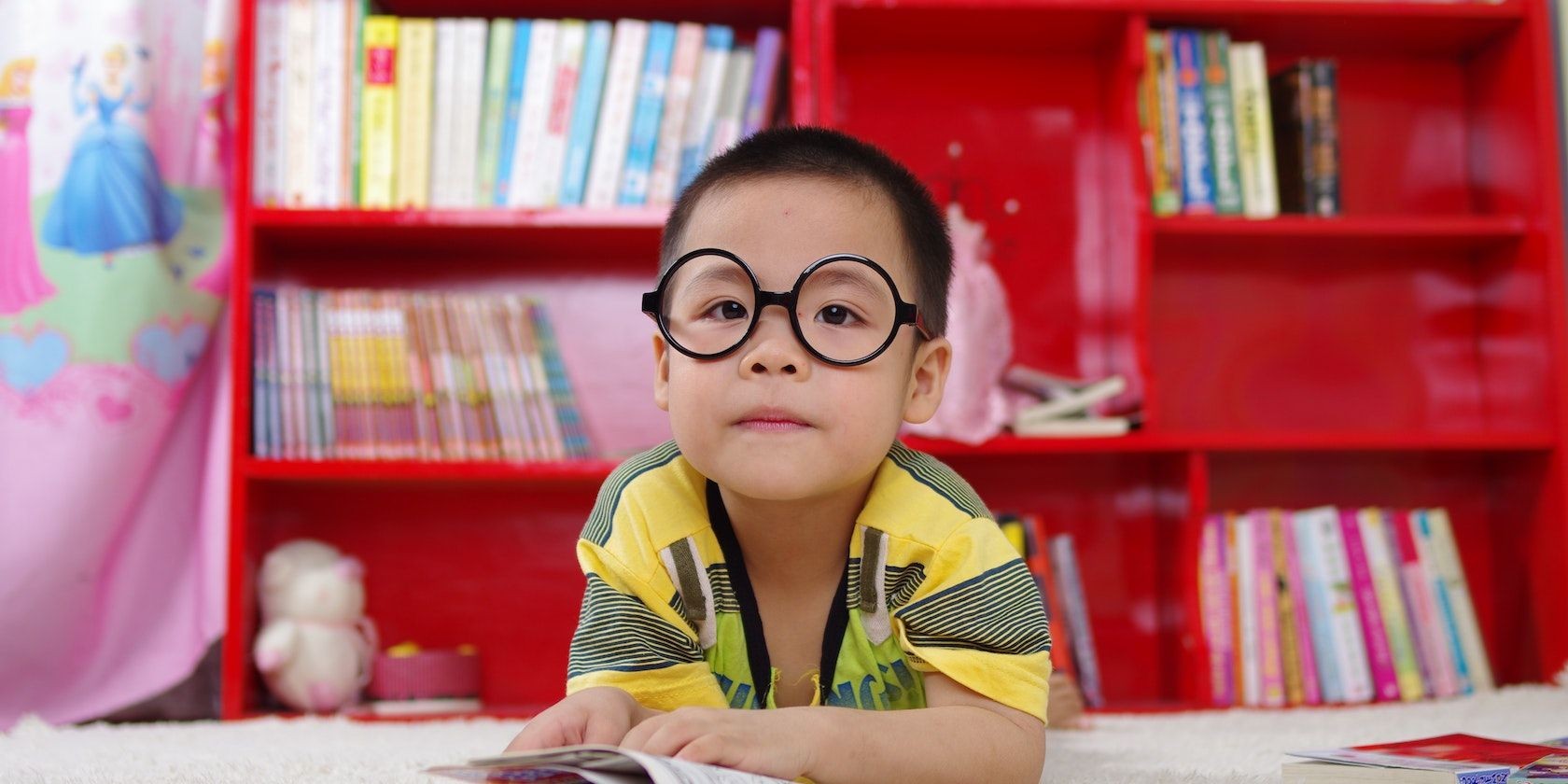 Trẻ em đeo kính quá khổ ngồi trong lớp học với kệ sách phía sau